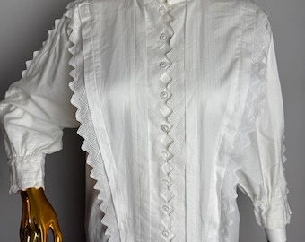 Pode incluir: Blusa branca de algodão com gola alta e fecho de botões. A blusa tem uma renda festonada em volta do colarinho, punhos e bainha.