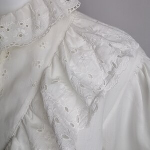 Puede incluir: Blusa blanca de algod&oacute;n con cuello de volantes y ribete de encaje de ojal. El encaje es un delicado patr&oacute;n de peque&ntilde;as flores y c&iacute;rculos.