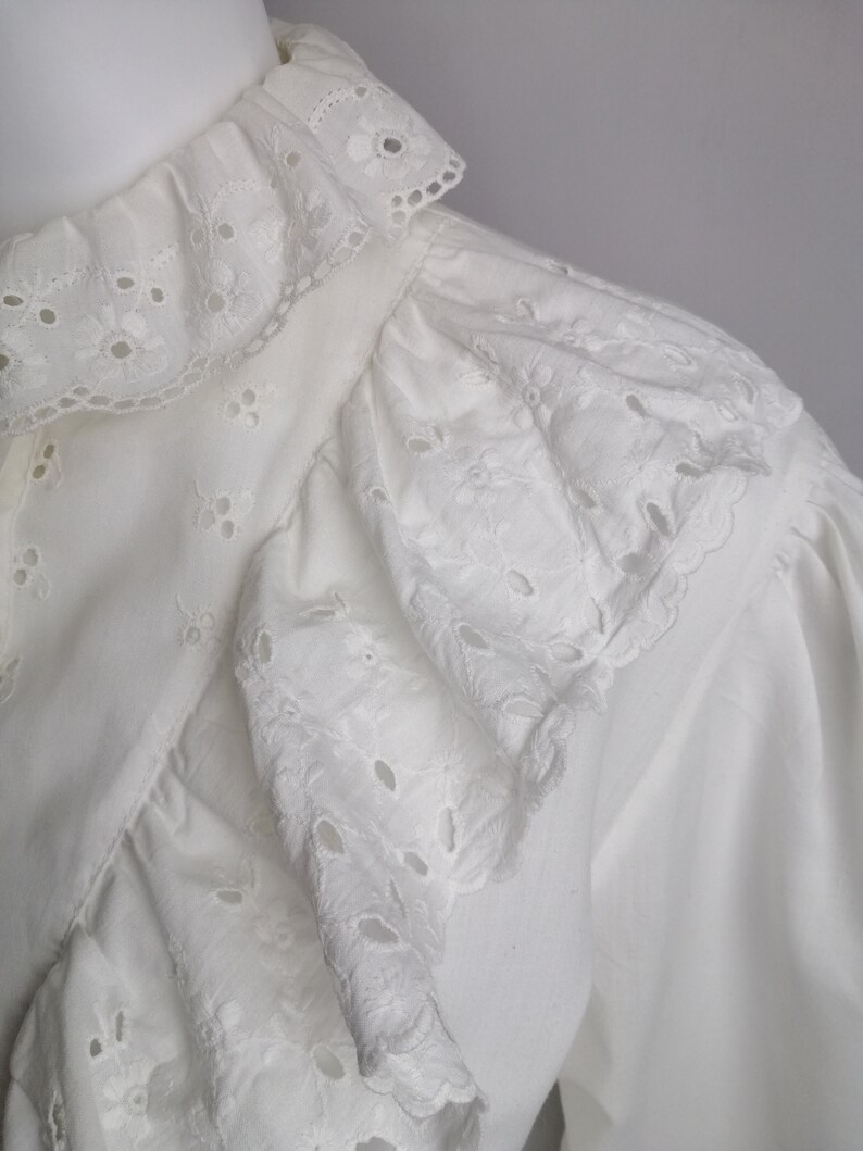 Puede incluir: Blusa blanca de algod&oacute;n con cuello de volantes y ribete de encaje de ojal. El encaje es un delicado patr&oacute;n de peque&ntilde;as flores y c&iacute;rculos.