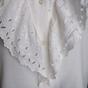Puede incluir: Una blusa blanca con un gran cuello con volantes con delicados detalles de encaje de ojal.