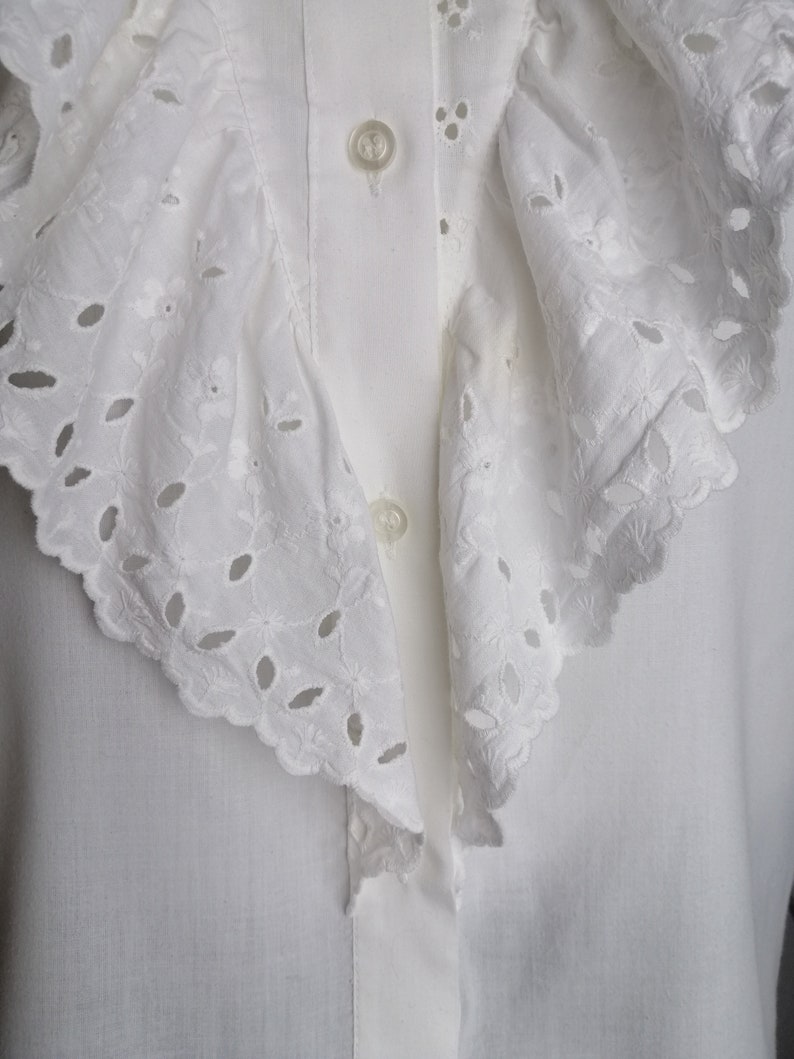 Puede incluir: Una blusa blanca con un gran cuello con volantes con delicados detalles de encaje de ojal.