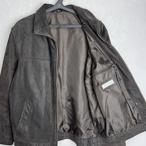 Puede incluir: Chaqueta de cuero marr&oacute;n oscuro con cierre de cremallera y cuello alzado. La chaqueta tiene dos bolsillos delanteros y un forro interior satinado. La chaqueta est&aacute; colgada de una percha negra.