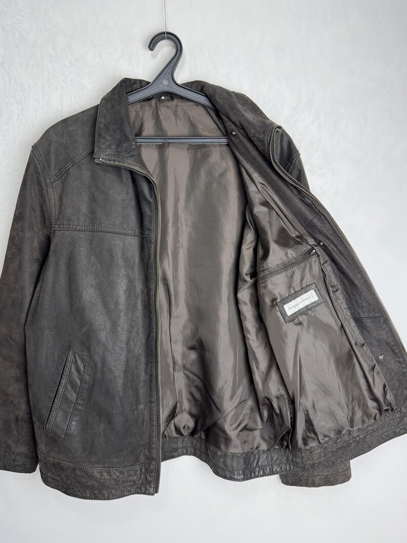Puede incluir: Chaqueta de cuero marr&oacute;n oscuro con cierre de cremallera y cuello alzado. La chaqueta tiene dos bolsillos delanteros y un forro interior satinado. La chaqueta est&aacute; colgada de una percha negra.