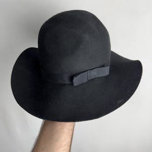 Puede incluir: Un sombrero fedora de fieltro negro con ala ancha y una cinta negra atada en un lazo en la parte delantera.