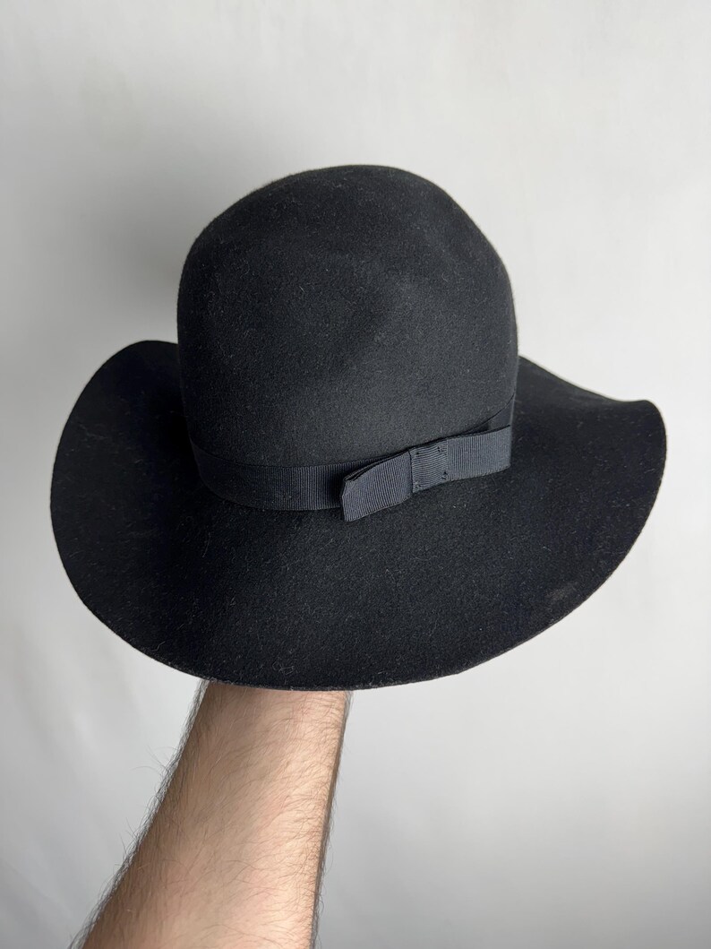 Puede incluir: Un sombrero fedora de fieltro negro con ala ancha y una cinta negra atada en un lazo en la parte delantera.