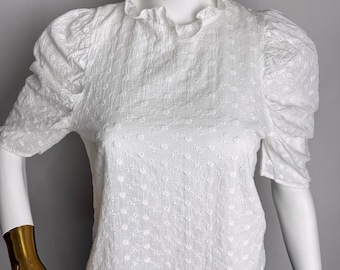 Pode incluir: Blusa branca de algodão com gola franzida e mangas curtas bufantes. A blusa tem um delicado padrão floral e é exibida em um manequim. O manequim está usando uma saia azul.
