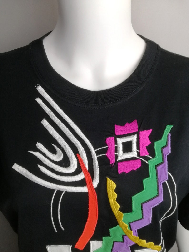 May include: Black t-shirt with a colourful abstract design featuring white, red, green, yellow, and purple embroidery.