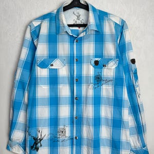 May include: A long-sleeve button-down shirt featuring a bright blue and white plaid pattern. The shirt includes two chest pockets, a collar, and black buttons. It is decorated with black ink-style graphics of a deer and postal stamps.