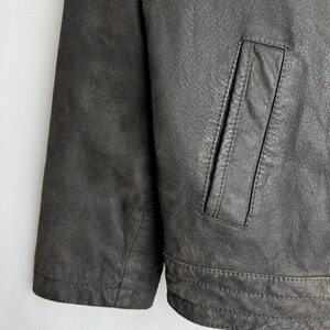 Puede incluir: Primer plano de una chaqueta de cuero gris oscuro. La chaqueta presenta una superficie texturizada, un solo bolsillo y detalles de costura visibles. La manga y el dobladillo inferior son visibles.