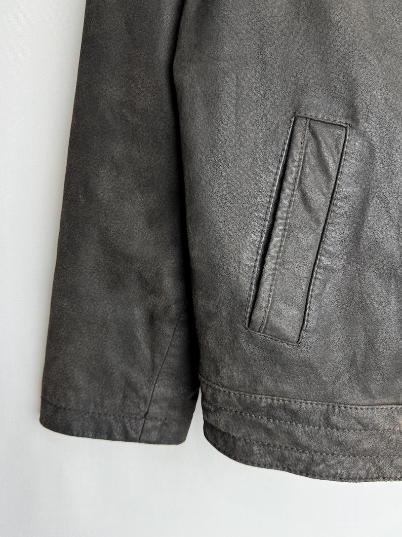 Puede incluir: Primer plano de una chaqueta de cuero gris oscuro. La chaqueta presenta una superficie texturizada, un solo bolsillo y detalles de costura visibles. La manga y el dobladillo inferior son visibles.