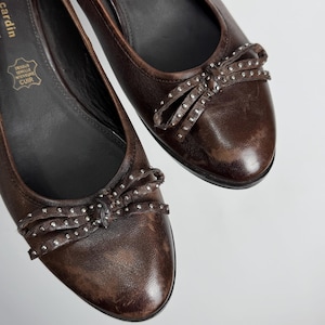 Puede incluir: Un par de bailarinas de cuero marr&oacute;n. Cada zapato presenta un lazo adornado con peque&ntilde;os tachuelas plateadas. Los zapatos tienen una punta redonda y un tac&oacute;n bajo. El logotipo de Pierre Cardin es visible.