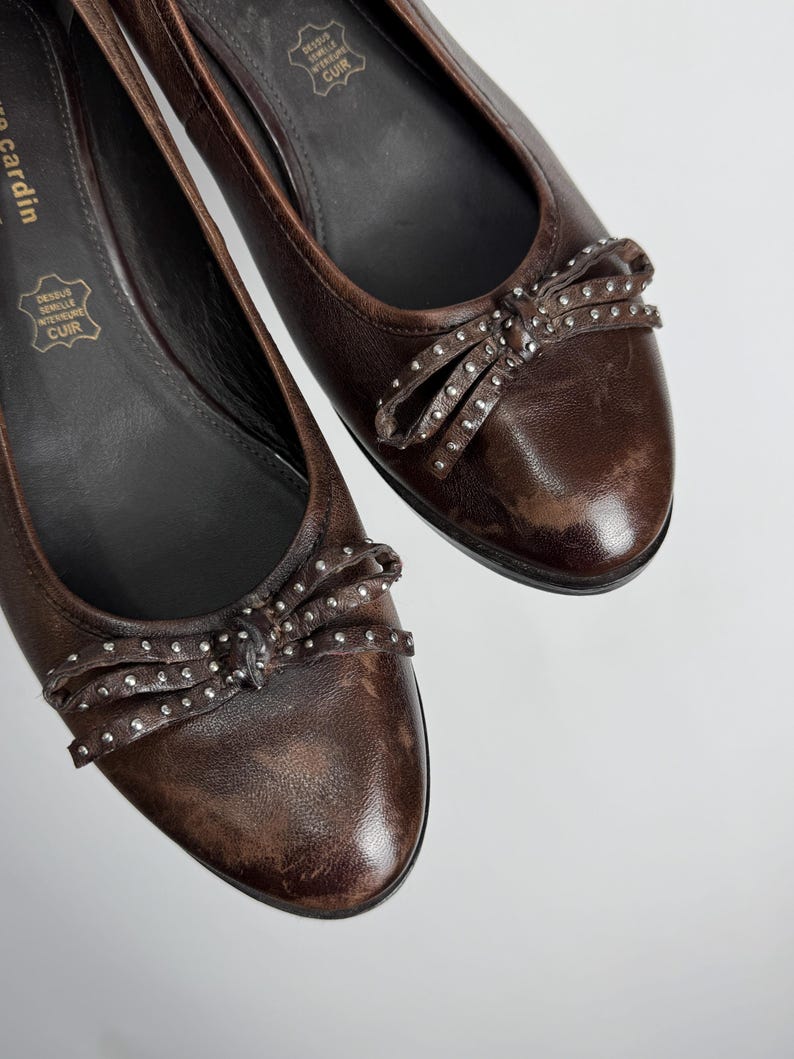 Puede incluir: Un par de bailarinas de cuero marr&oacute;n. Cada zapato presenta un lazo adornado con peque&ntilde;os tachuelas plateadas. Los zapatos tienen una punta redonda y un tac&oacute;n bajo. El logotipo de Pierre Cardin es visible.