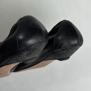 Puede incluir: Par de bailarinas de cuero negro. Los zapatos tienen una punta redondeada y un peque&ntilde;o tac&oacute;n de cu&ntilde;a. Las suelas muestran signos de desgaste. El cuero parece ligeramente arrugado. Los zapatos son probablemente usados.