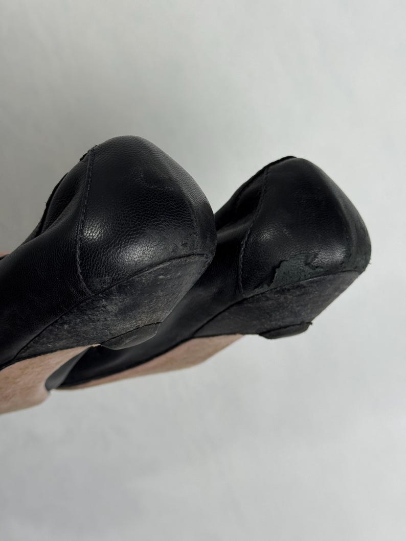 Puede incluir: Par de bailarinas de cuero negro. Los zapatos tienen una punta redondeada y un peque&ntilde;o tac&oacute;n de cu&ntilde;a. Las suelas muestran signos de desgaste. El cuero parece ligeramente arrugado. Los zapatos son probablemente usados.