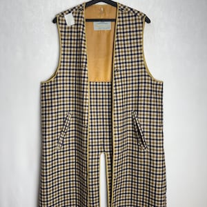 Könnte beinhalten: Eine lange, ärmellose Weste mit einem karierten Muster in Marineblau, Beige und Weiß. Die Weste hat ein beiges Futter und eine beige Paspelierung sowie zwei Vordertaschen. Sie hängt an einem schwarzen Kleiderbügel.