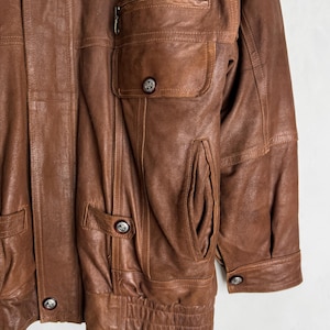 May include: A brown leather jacket with multiple pockets and silver-toned buttons. The jacket has a zipper pocket on the chest and a ribbed hem. The leather appears worn, giving it a vintage look.