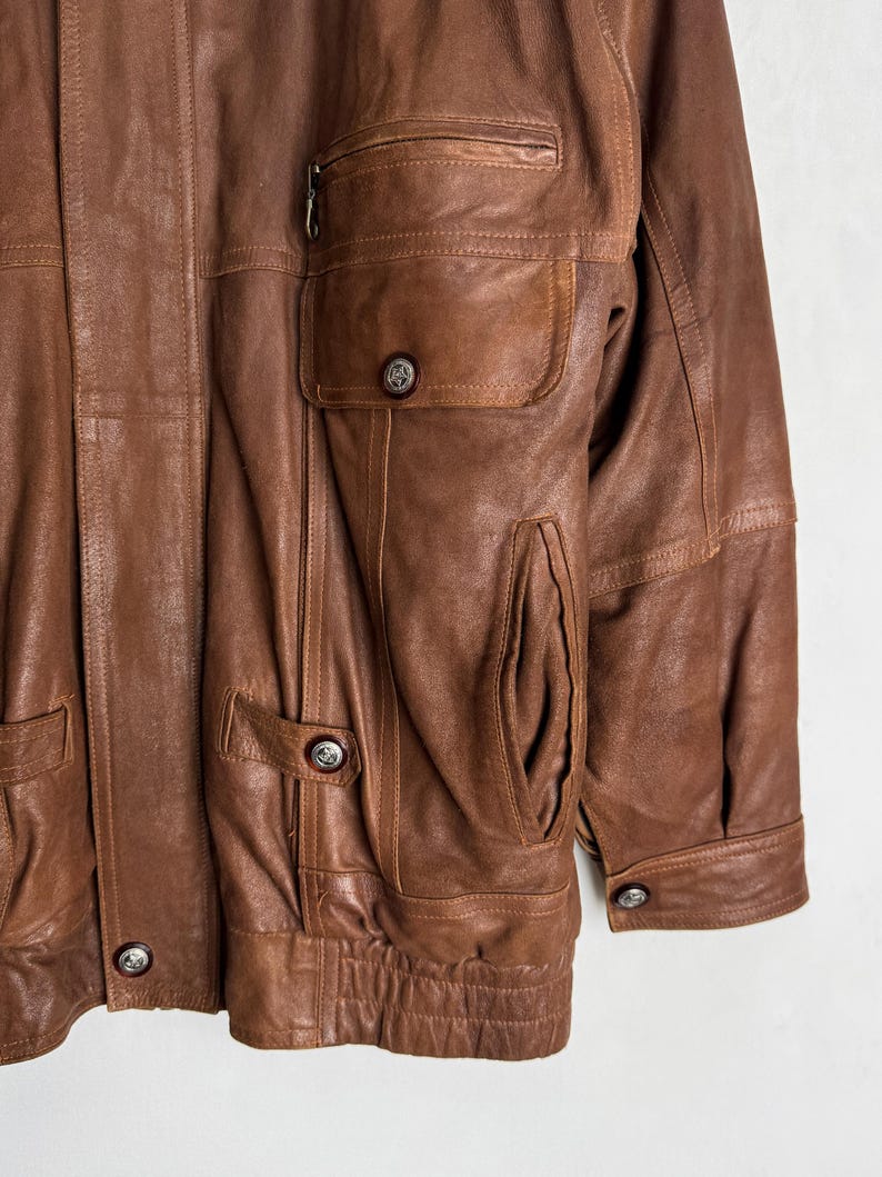 May include: A brown leather jacket with multiple pockets and silver-toned buttons. The jacket has a zipper pocket on the chest and a ribbed hem. The leather appears worn, giving it a vintage look.