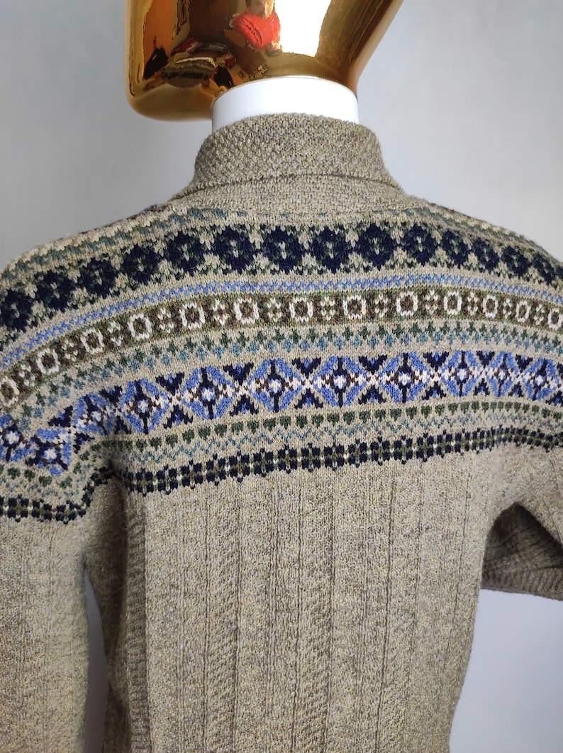 May include: A tan-colored sweater with a patterned design across the shoulders and upper back. The pattern includes blue, green, and white geometric shapes. The sweater has a ribbed texture on the back.