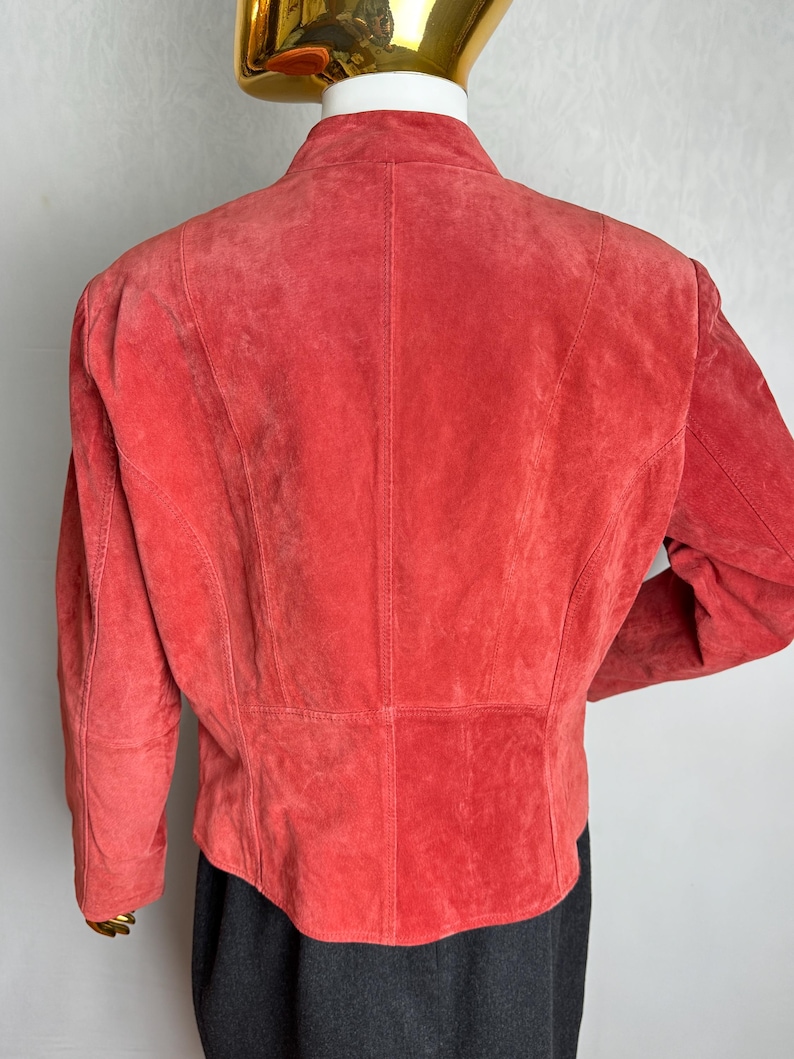 Puede incluir: Chaqueta de ante color coral con cuello alzado y mangas largas. La parte trasera de la chaqueta presenta costuras verticales y un corte entallado. La chaqueta se exhibe en un maniqu&iacute;.