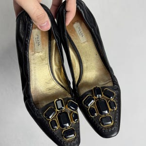 Puede incluir: Un par de bailarinas Prada de cuero negro. Los zapatos tienen una plantilla dorada y est&aacute;n adornados con joyas rectangulares negras engastadas en metal dorado. El logotipo de Prada es visible en la plantilla.