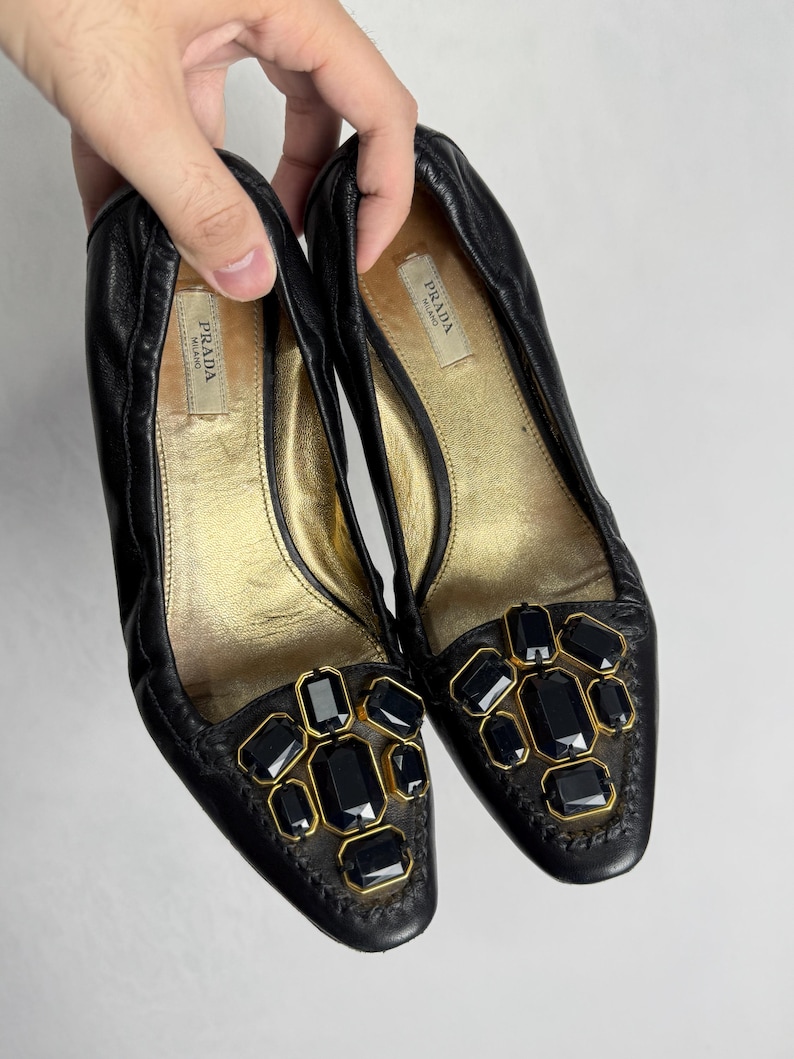 Puede incluir: Un par de bailarinas Prada de cuero negro. Los zapatos tienen una plantilla dorada y est&aacute;n adornados con joyas rectangulares negras engastadas en metal dorado. El logotipo de Prada es visible en la plantilla.