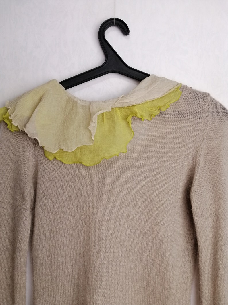 Puede incluir: Un su&eacute;ter de punto beige con un cuello de volantes en tonos amarillos y blancos.
