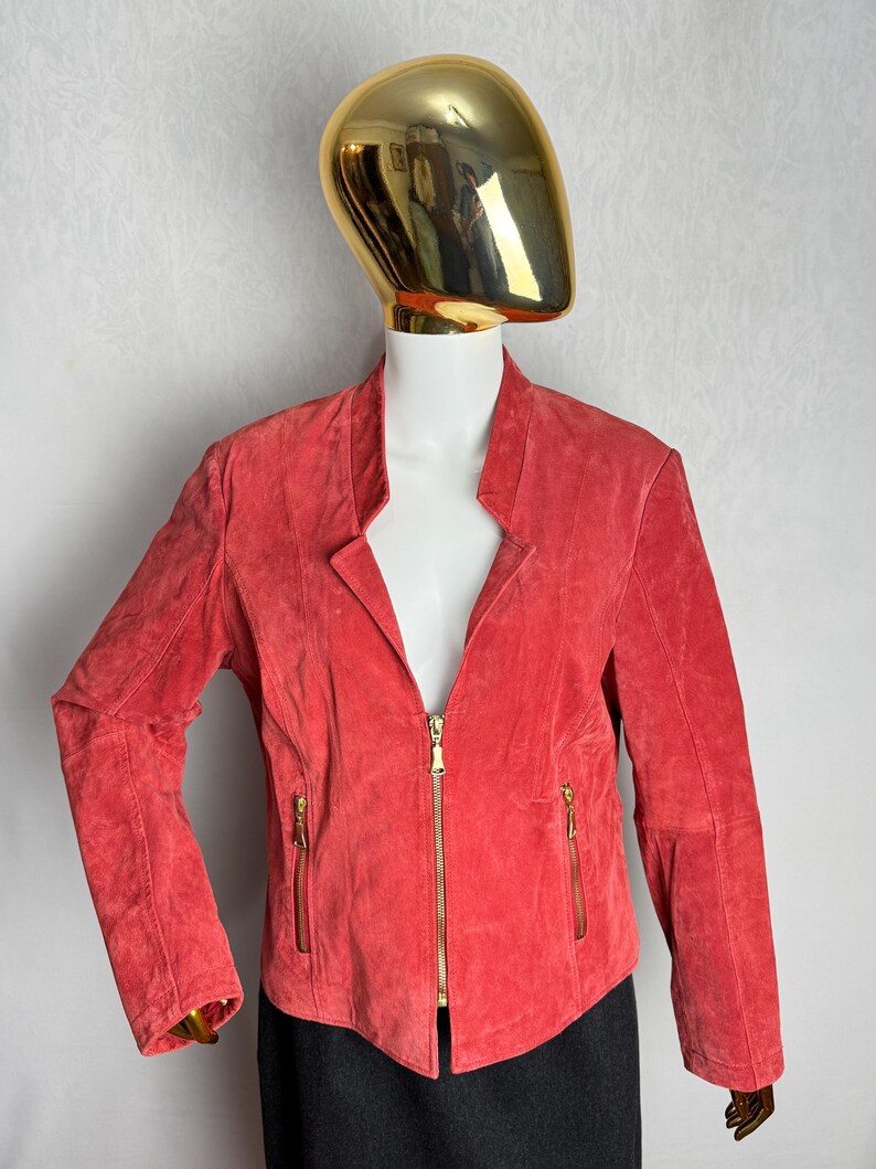 Puede incluir: Chaqueta de ante color coral con cuello alzado y cremallera completa. La chaqueta tiene dos bolsillos con cremallera y mangas largas. La chaqueta se exhibe en un maniqu&iacute;.