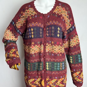 Op de afbeelding: Een gebreid vest met lange mouwen en knoopsluiting in een diepe bordeauxrode kleur. Het kledingstuk heeft een veelkleurig geometrisch patroon in tinten mosterdgeel, bosgroen, marineblauw en roze, met een geschulpte rand langs de halslijn.