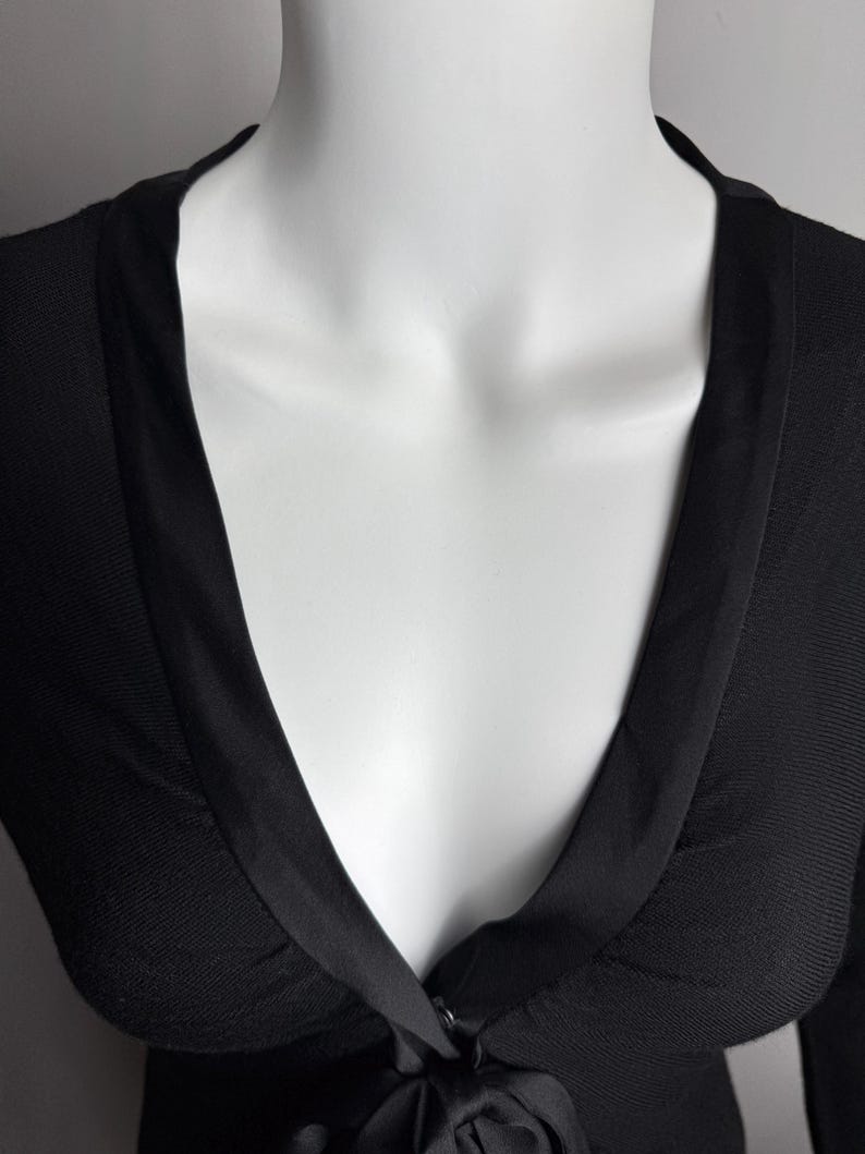K&ouml;nnte beinhalten: Ein schwarzer Strickcardigan mit tiefem V-Ausschnitt und Satinbesatz. Der Cardigan hat einen Knopfverschluss und eine Schleife an der Taille.