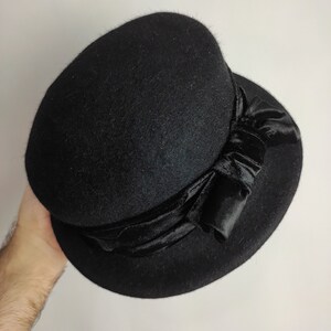 May include: A black cloche hat with a rounded crown and a wide brim. The hat features a black velvet ribbon tied in a bow on the side. The hat is made of a soft, felt-like material.