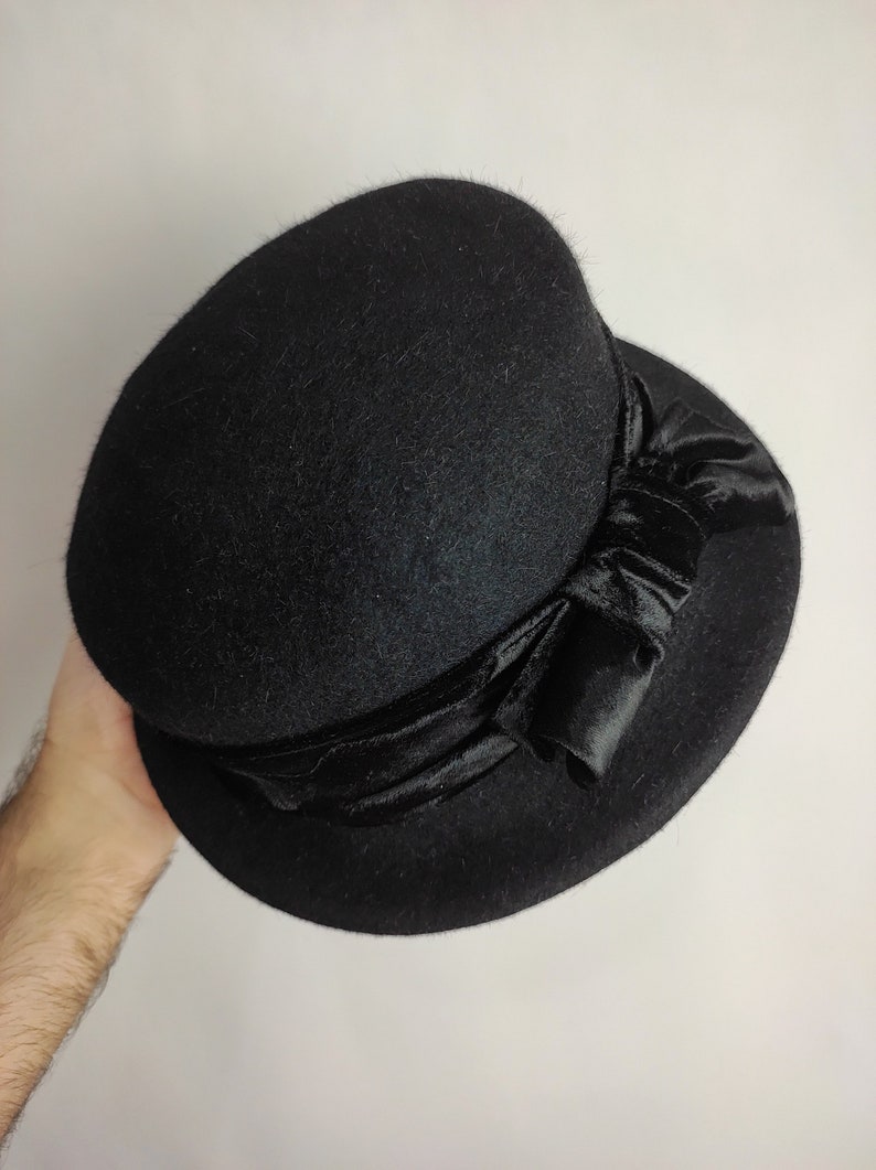 May include: A black cloche hat with a rounded crown and a wide brim. The hat features a black velvet ribbon tied in a bow on the side. The hat is made of a soft, felt-like material.