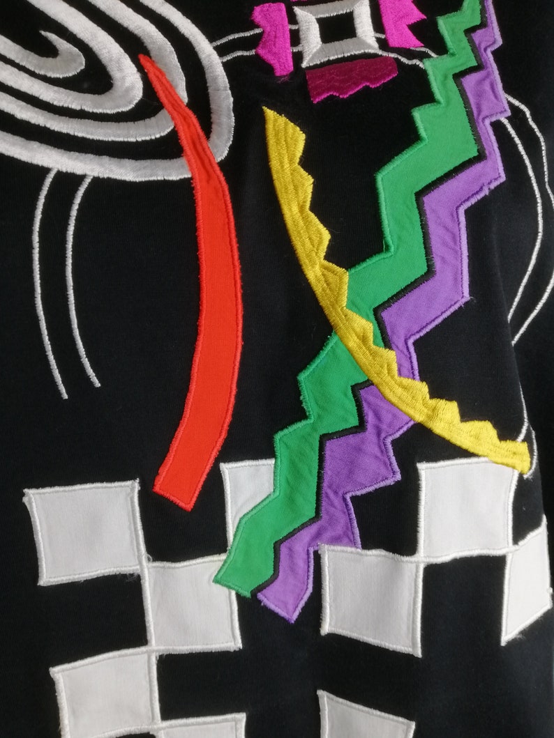 May include: Black fabric with colourful embroidered geometric shapes. The shapes include a spiral, a zig-zag, a curved line, and a chequered pattern.