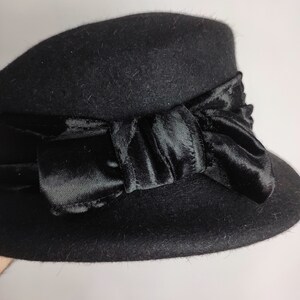 May include: A black cloche hat with a velvet bow. The hat is made of a soft, felt-like material and features a wide brim. The bow is a dark, rich black and adds a touch of elegance to the hat. This hat is a stylish accessory.