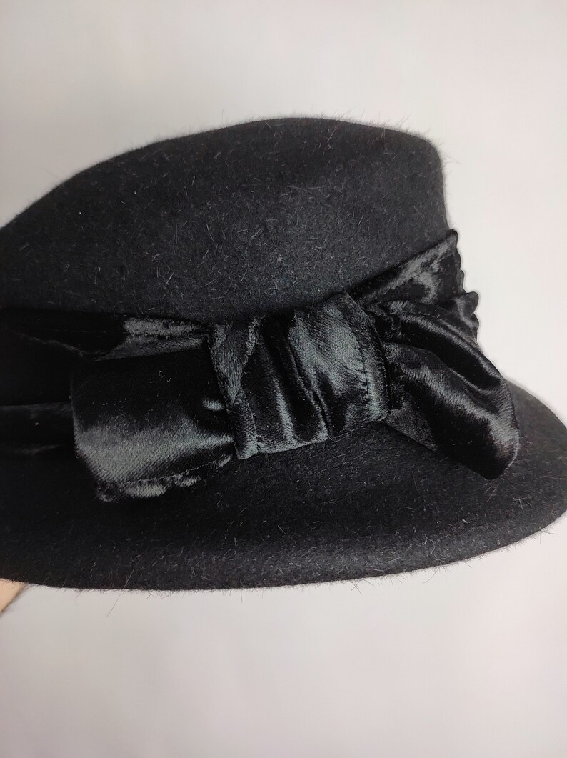 May include: A black cloche hat with a velvet bow. The hat is made of a soft, felt-like material and features a wide brim. The bow is a dark, rich black and adds a touch of elegance to the hat. This hat is a stylish accessory.
