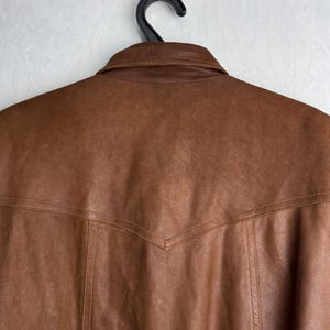 May include: Close-up of a brown leather jacket hanging on a black hanger. The jacket has a collar and detailed stitching along the seams. The leather has a slightly worn appearance.