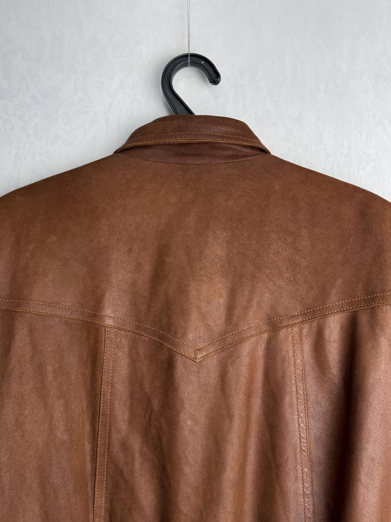 May include: Close-up of a brown leather jacket hanging on a black hanger. The jacket has a collar and detailed stitching along the seams. The leather has a slightly worn appearance.