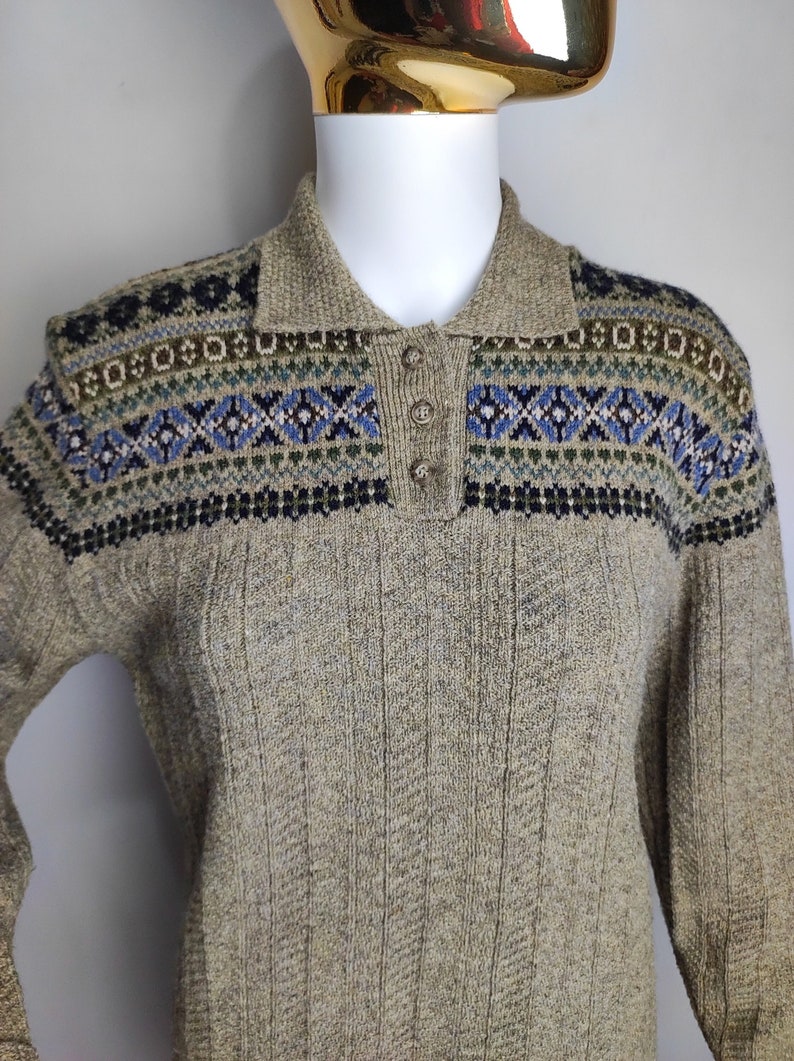 May include: A tan sweater with a collared neckline and three buttons. The yoke features a geometric pattern in blue, green, and black. The sweater has a ribbed texture.