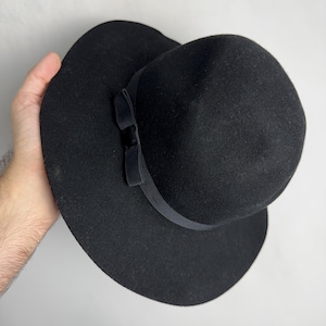 Puede incluir: Un sombrero fedora de fieltro negro con ala ancha y un lazo de cinta negro.