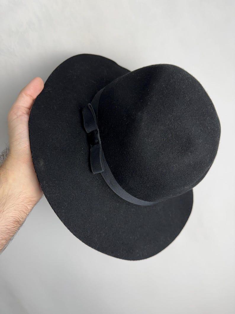 Puede incluir: Un sombrero fedora de fieltro negro con ala ancha y un lazo de cinta negro.