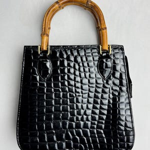 May include: A black handbag with a glossy, embossed crocodile skin texture. The bag features a rounded bamboo handle attached with gold-toned metal hardware. The structure is square-shaped with rounded bottom corners and a smooth, shiny finish.