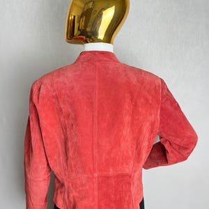 Puede incluir: Chaqueta de ante color coral con cuello alzado. La vista trasera muestra las costuras a medida y un dobladillo ligeramente curvado. La chaqueta se exhibe en un maniqu&iacute; con cabeza dorada.