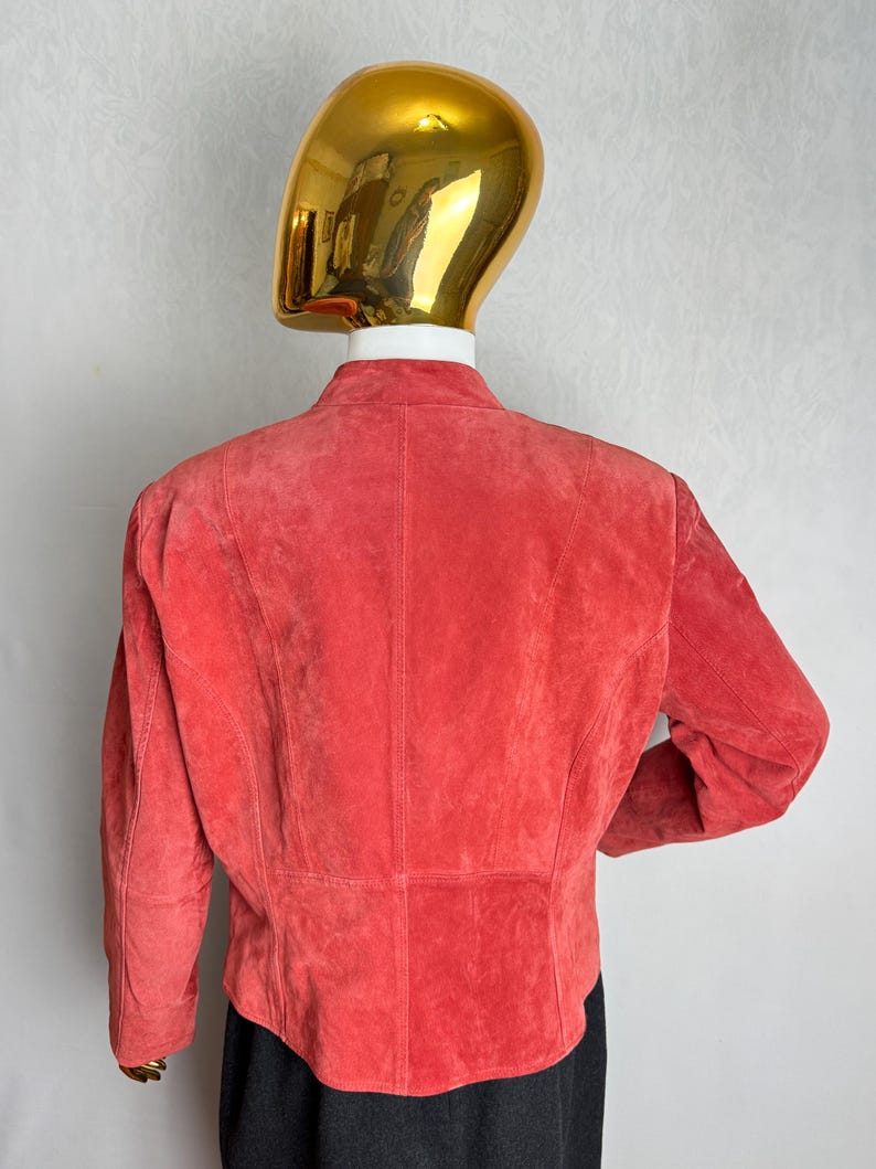 Puede incluir: Chaqueta de ante color coral con cuello alzado. La vista trasera muestra las costuras a medida y un dobladillo ligeramente curvado. La chaqueta se exhibe en un maniqu&iacute; con cabeza dorada.
