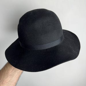 Puede incluir: Un sombrero fedora de fieltro negro con ala ancha y una cinta negra.
