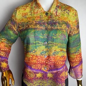 May include: A long-sleeved, button-up shirt with a colourful, abstract print. The shirt has a ruffled hem and a collar. The print features shades of green, orange, yellow, and purple.