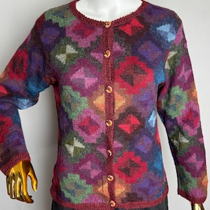 Peut inclure: Cardigan coloré à motifs avec un col rond et des manches longues. Le pull à boutons présente un motif géométrique dans les tons de rouge, violet, vert et bleu. La bordure et les boutons sont de couleur rouge complémentaire.