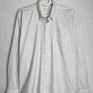 Puede incluir: Camisa blanca abotonada con un patrón de cuadrícula en color gris claro. La camisa de manga larga tiene un bolsillo en el pecho y está colgada de una percha blanca. La camisa está hecha de una tela ligera.