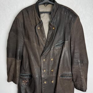 May include: A vintage brown leather jacket with a double-breasted design. The jacket features gold-colored buttons and a classic lapel. The leather has a distressed appearance, adding to its vintage appeal. The jacket is hanging on a black hanger.
