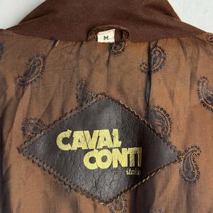 May include: Close-up of the inside of a brown jacket. The lining is a paisley pattern in shades of brown and blue. A brown leather patch with the words "CAVAL CONTI" in gold lettering is sewn into the lining. The jacket's collar is a darker shade of brown.