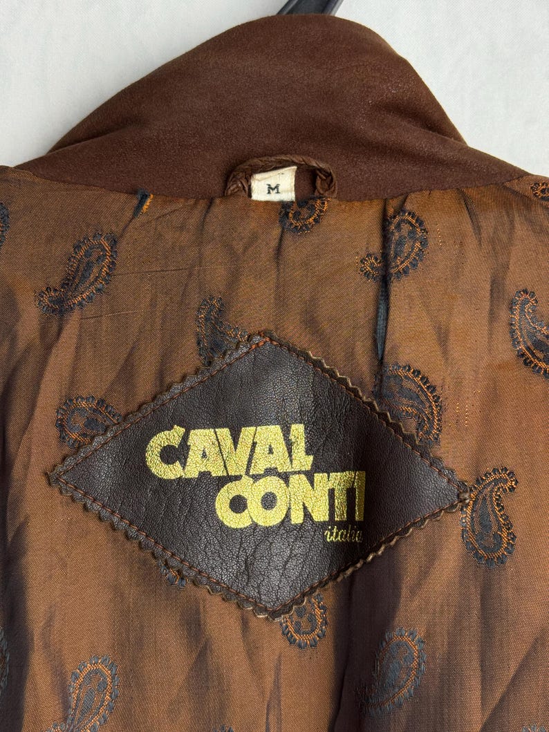 May include: Close-up of the inside of a brown jacket. The lining is a paisley pattern in shades of brown and blue. A brown leather patch with the words "CAVAL CONTI" in gold lettering is sewn into the lining. The jacket's collar is a darker shade of brown.