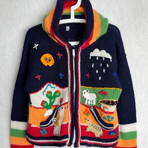 May include: A navy blue zip-up cardigan with colourful embroidered designs. The cardigan features a rainbow-striped collar and cuffs, and various appliqued images including a sun, cloud, cactus, and animals. The cardigan is hanging on a white hanger.